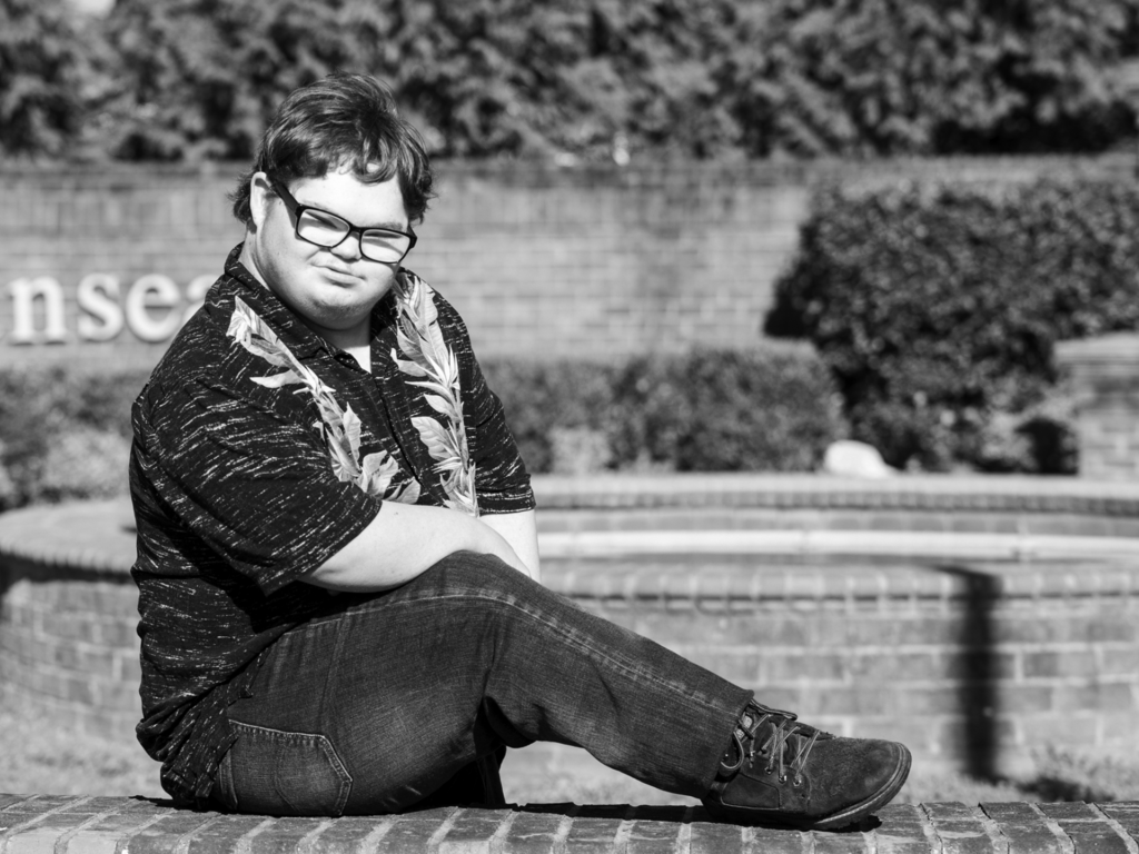 Black and white photo of young adult male sitting on brick ledge in front of brick wall and bushes
