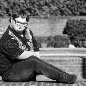 Black and white photo of young adult male sitting on brick ledge in front of brick wall and bushes