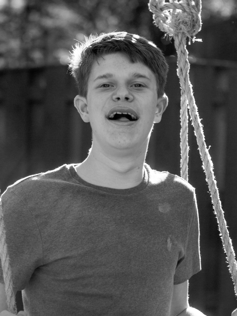 Black and white photo of a young adult male sitting on a rope swing