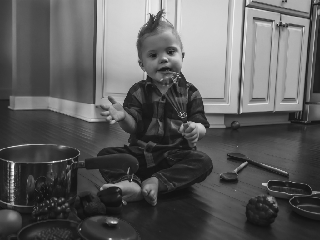 Black and white photo of infant holding a whisk surrounded by kitchen utensils