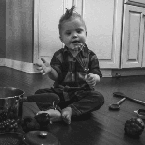 Black and white photo of infant holding a whisk surrounded by kitchen utensils