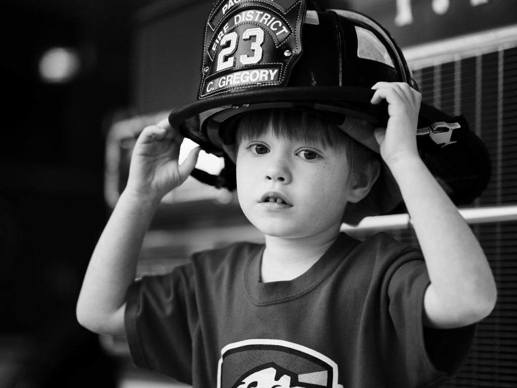 Black and white photo of male child wearing a fireman’s helmet