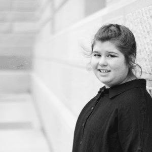 Black and white photo of young adult female leaning against wall in a dark button down shirt