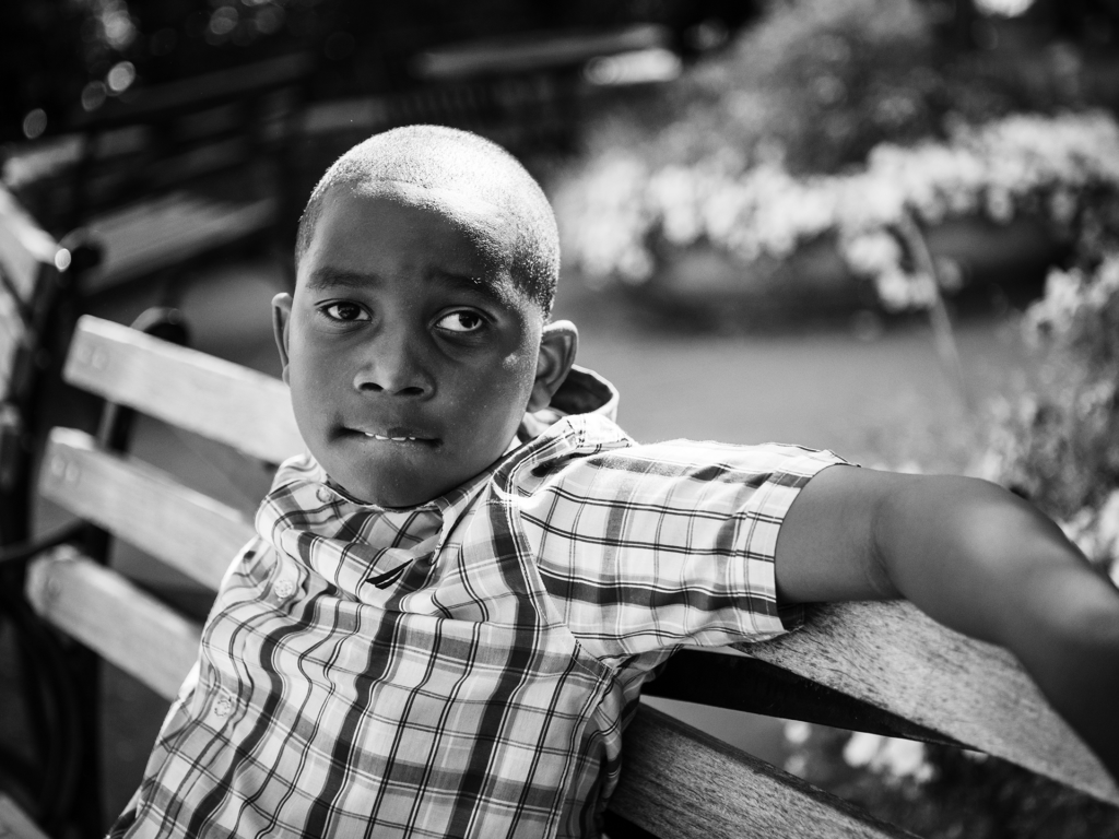 Black and white photo of male child sitting on a bench outside in a plaid shirt