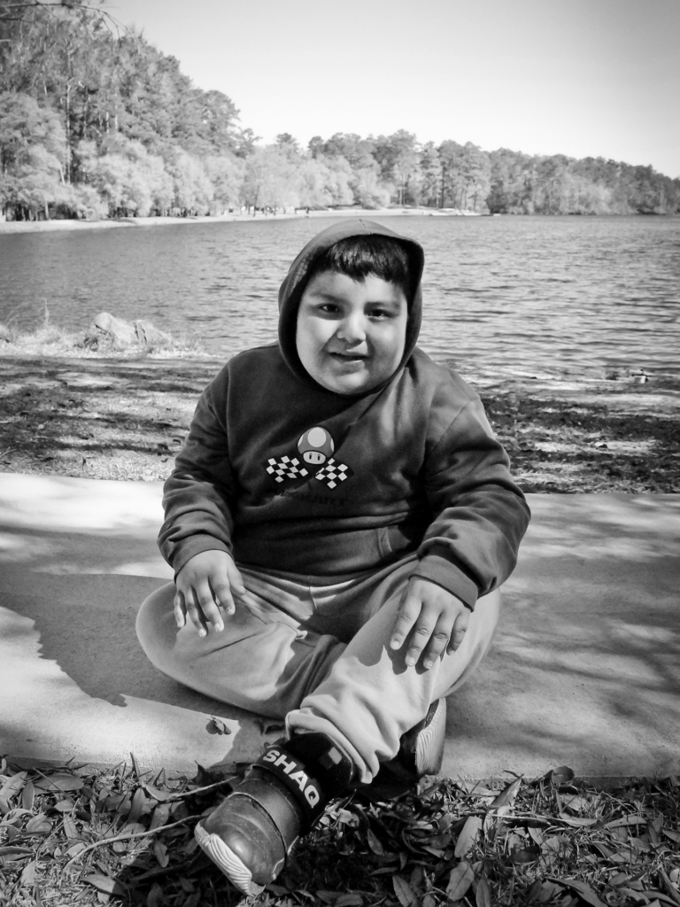 Black and white photo of male child sitting on a sidewalk in front of a lake