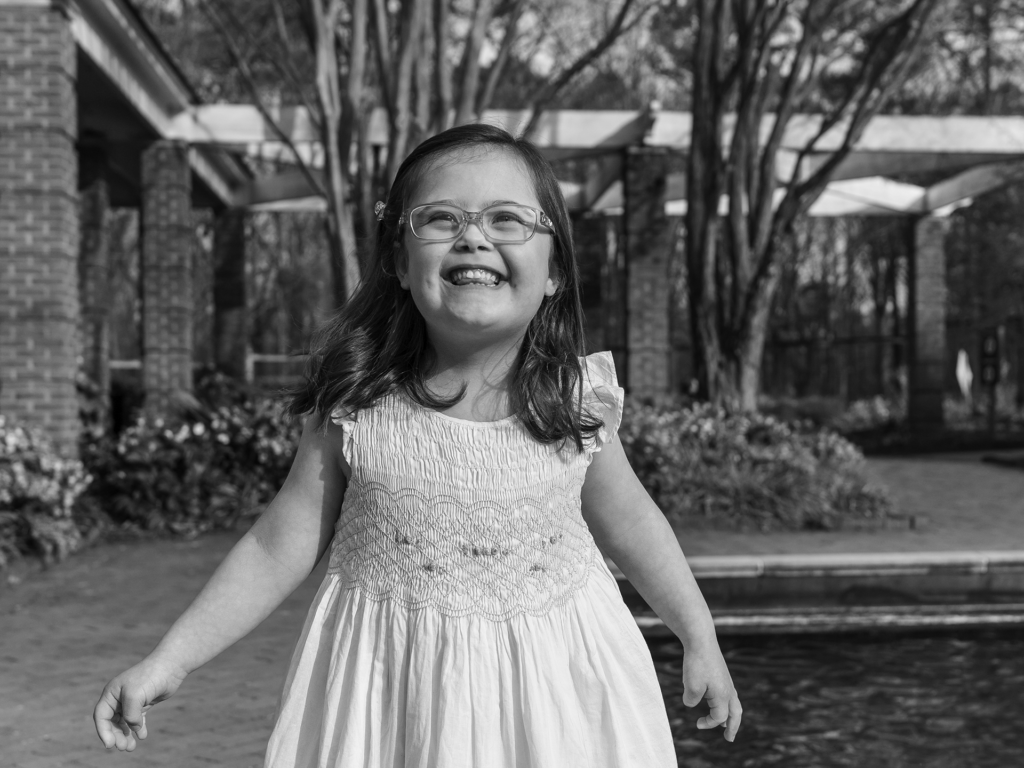 Black and white photo of female child smiling in front of trees and bushes beside a pool