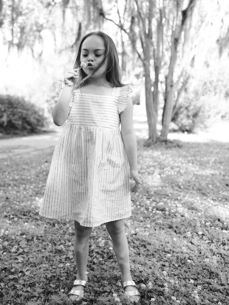 Black and white photo of female child blowing a dandelion outside in grass