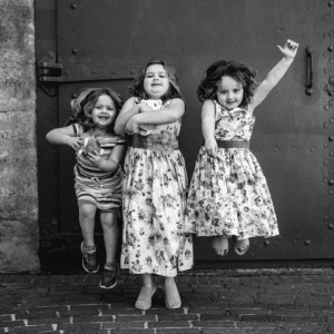 Black and white photo of three female children jumping in front of metal door