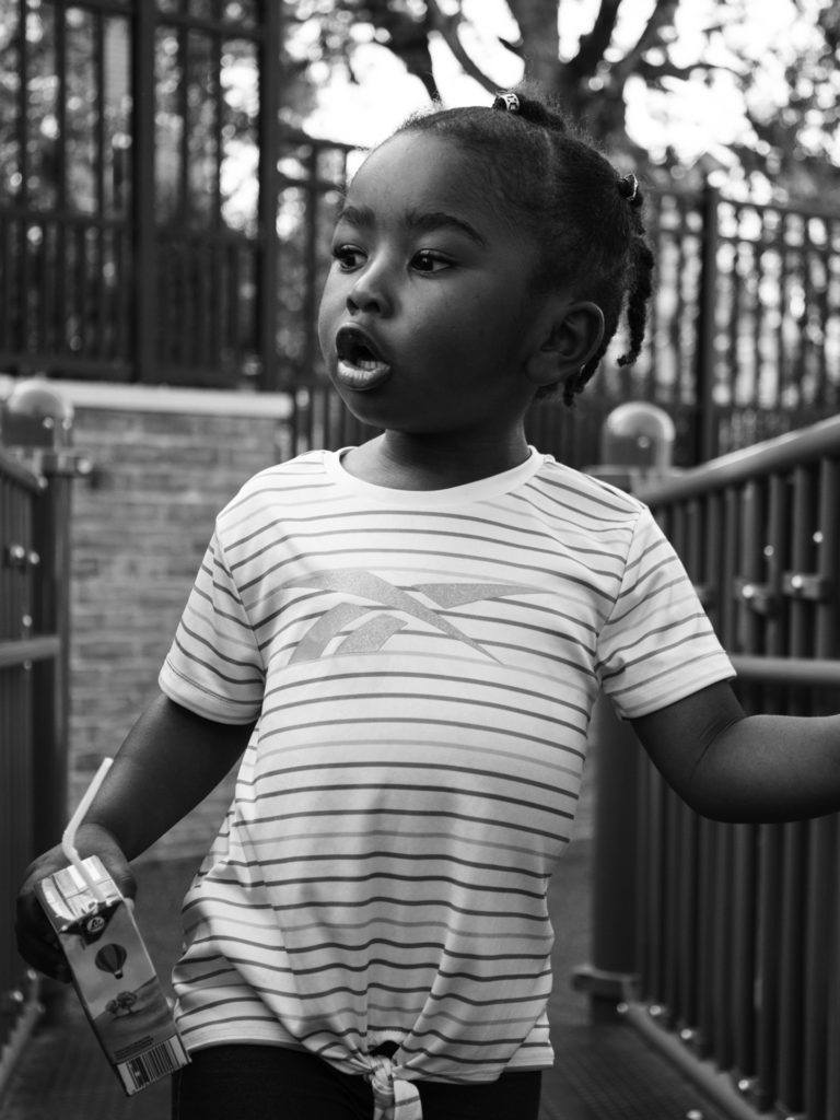 Black and white photo of female toddler walking across playground bridge with a juice
